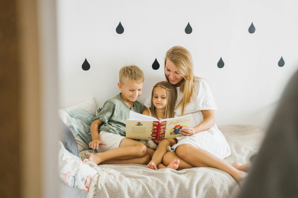 A joyful mother reads a storybook to her young children while sitting on a bed.
