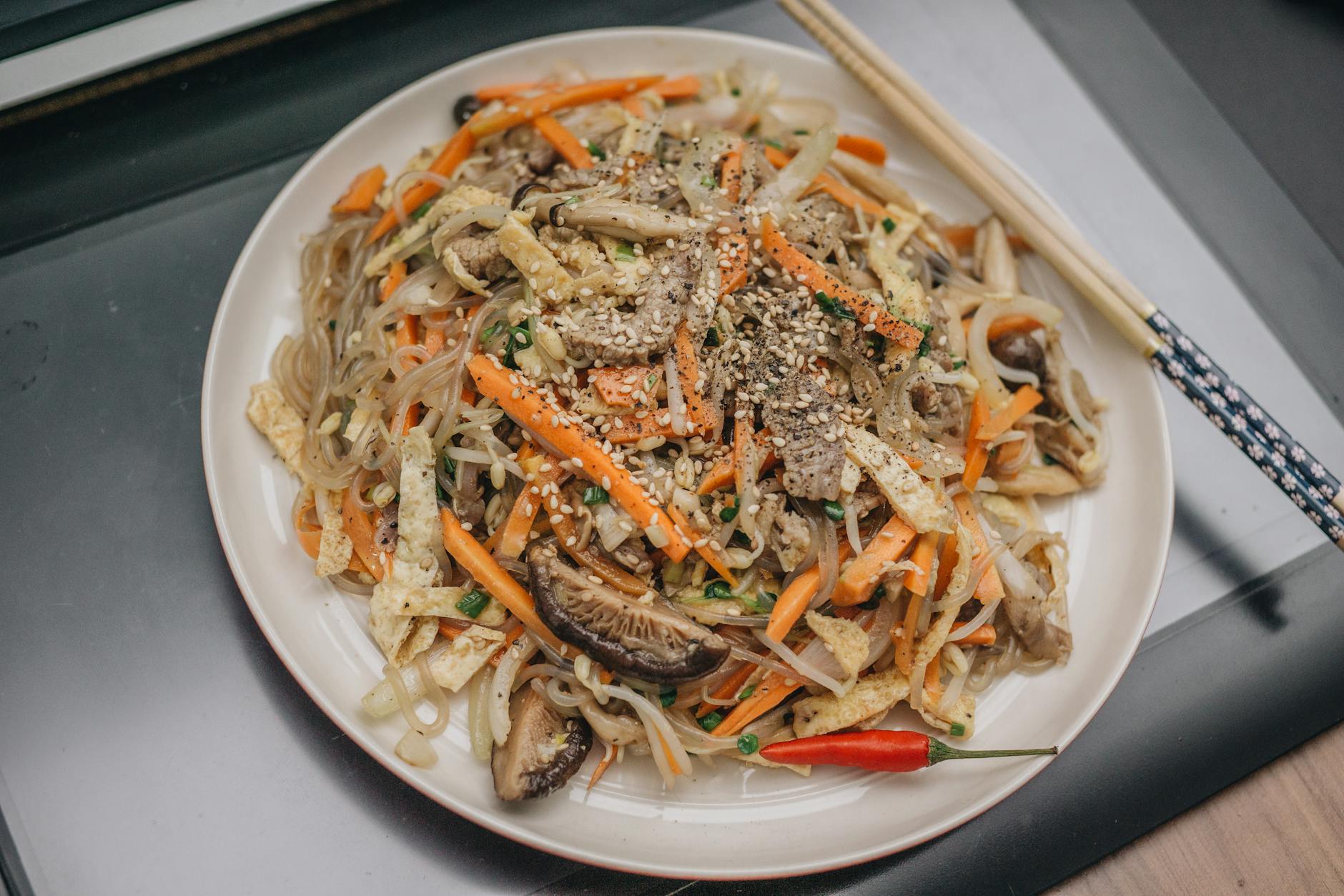Colorful stir-fried noodles with vegetables and sesame seeds, garnished with chili and served with chopsticks.